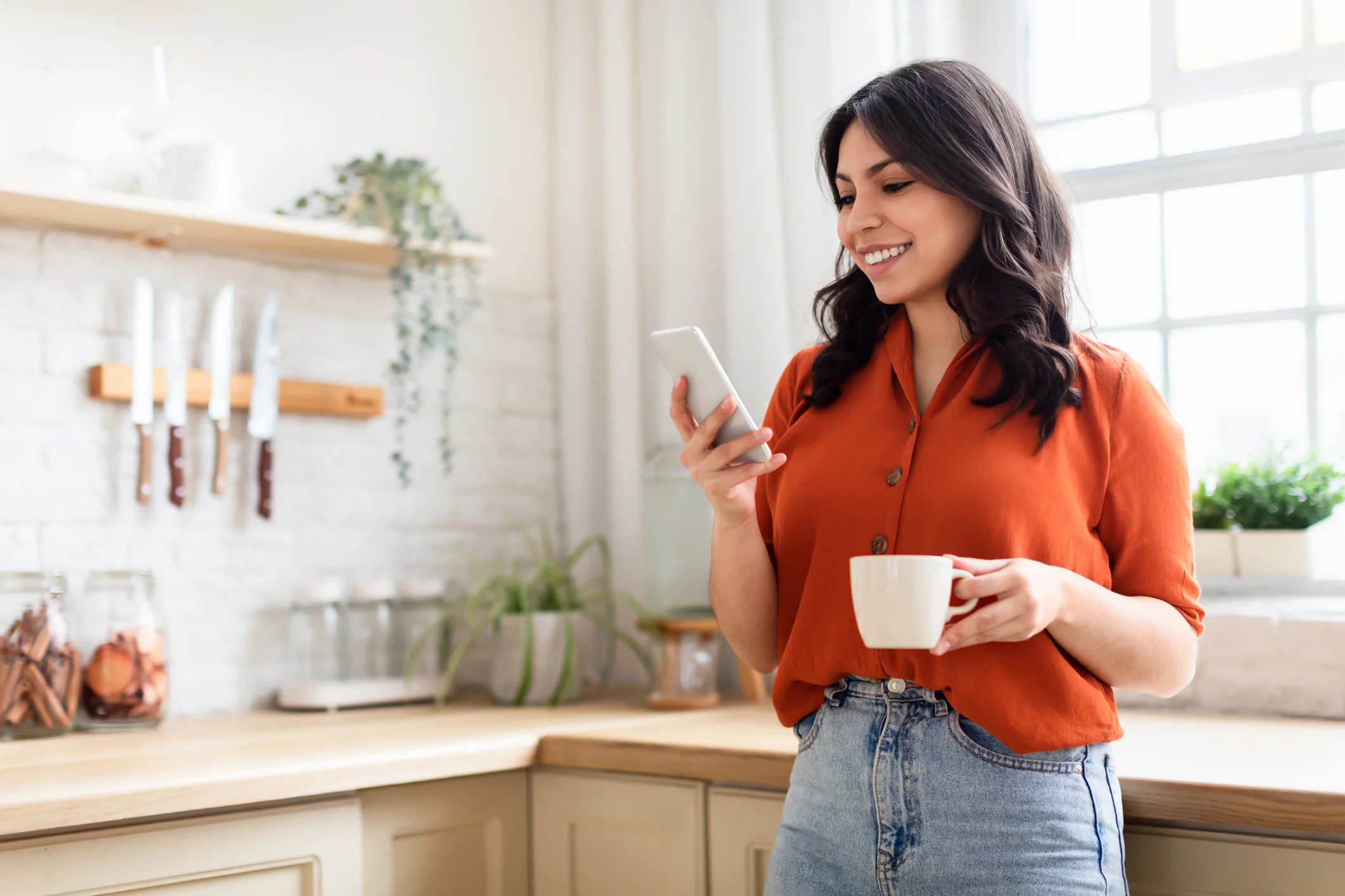 Lächelnde Person hält eine Tasse und schaut in einer Küche auf ihr Smartphone.