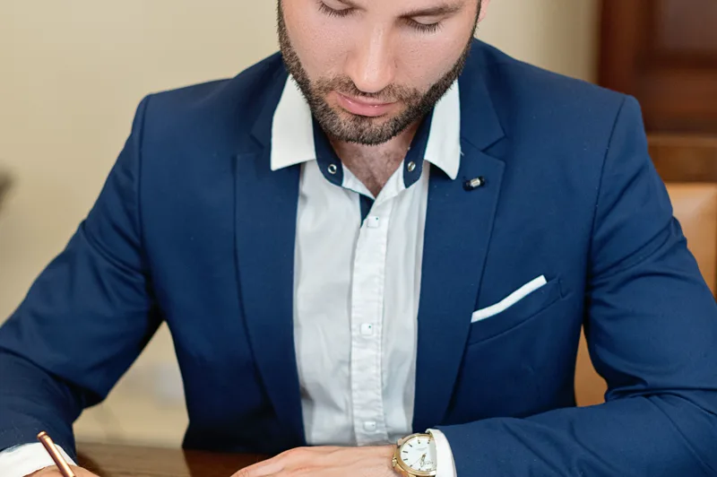 Mann im blauen Anzug liest und unterschreibt ein Dokument an seinem Schreibtisch.