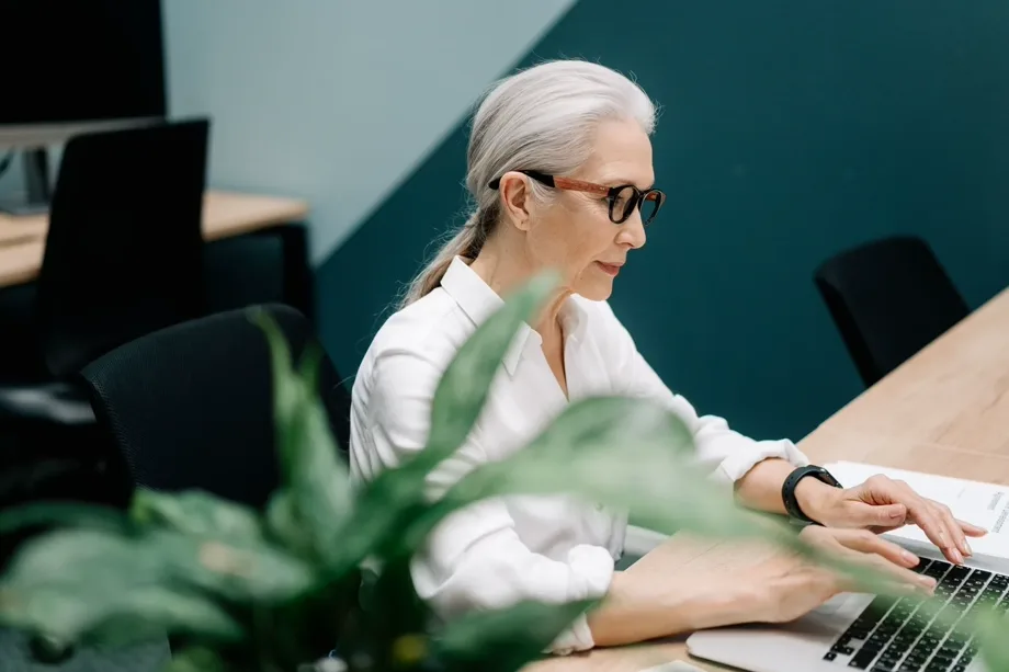 Una donna dai capelli grigi, in camicia bianca e occhiali, lavora al computer portatile in un ufficio, con una pianta in primo piano.