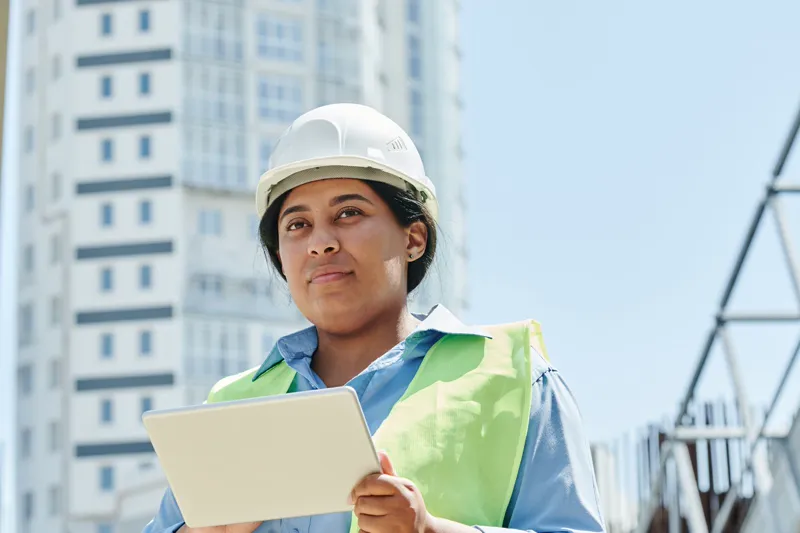Une ingénieure portant un casque de chantier consulte une tablette devant un immeuble en construction.