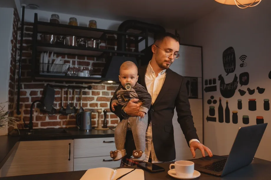Ein Vater im Anzugjackett hält sein Baby im Arm und arbeitet in seiner Küche am Laptop.